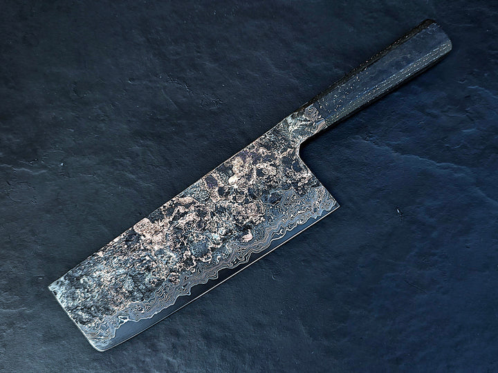 One-of-a-kind Cu-Ni-Mascus Nakiri knife by Leszek Sikon, showcasing 8120 layers of Damascus forged from Spicy White steel, O1 tool steel, copper, nickel, and brass. The blade features a rich, geological pattern with molten textures and wave-like lines, hand-forged over 100 hours with an S-profile for food release and finished with a custom jeans micarta handle embedded with copper and steel mesh.