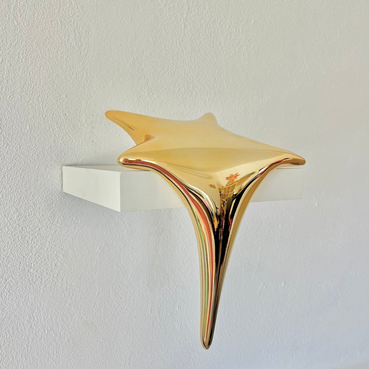 Polished gold-toned stainless steel sculpture in the form of a stylised shooting star, with a fluid, elongated spike melting over the edge of a white plinth or shelf.