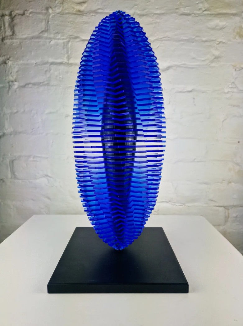 Sculptural artwork “Heliocentric Worlds” by Richard Slee, crafted from stacked layers of translucent neon blue acrylic forming an elongated, seed-like form. Mounted on a minimalist steel base, the piece emits a vivid blue glow, casting intricate shadows and reflecting ambient light to create a striking visual effect. Sleek, symmetrical, and futuristic in presence.