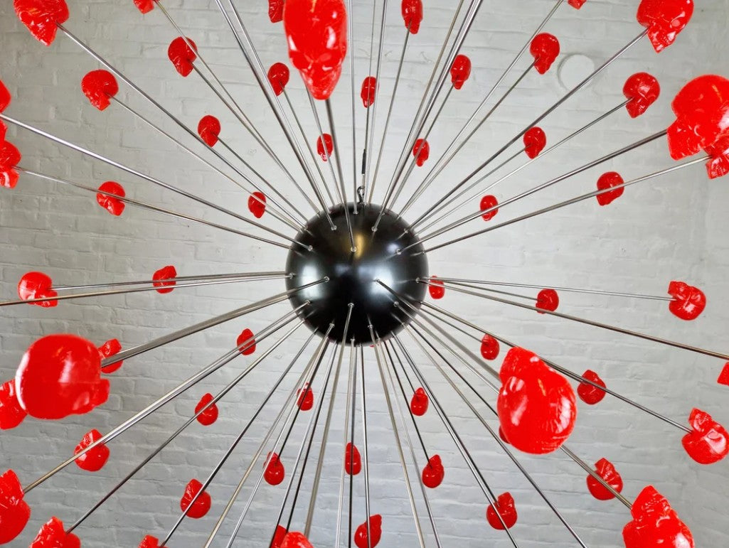 Large spherical hanging sculpture composed of a central black metallic orb with over 70 radiating stainless steel rods, each tipped with a glossy red sculpted skull. The skulls appear to orbit the sphere like a molecular explosion or stylised dandelion, creating a dramatic and surreal visual impact. Materials include stainless steel, polymorph and resin, with vibrant red gloss details and industrial metallic finishes.