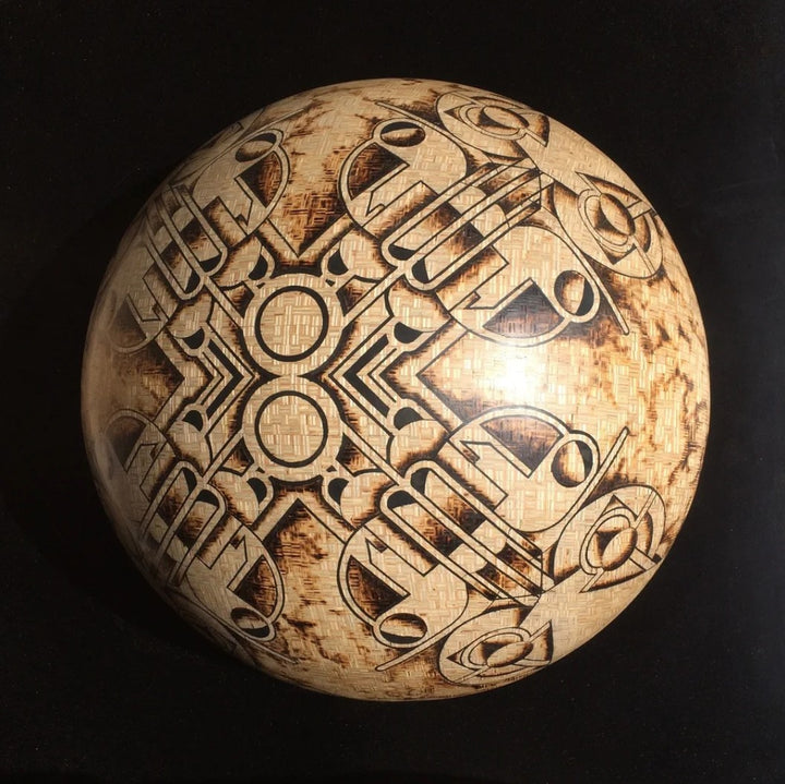 Intricately engraved hemispherical sculpture by Richard Slee titled “New Horizons Approaching.” Crafted from square 9mm plywood and shaded using a controlled burn technique, the surface features symmetrical, abstract motifs inlaid with black putty. The design has a radial, orbital quality with complex geometric lines and scorched textures, creating a striking contrast between natural wood tones and dark patterning.