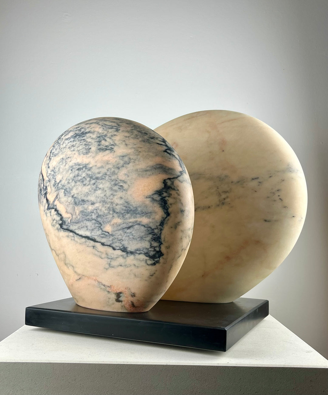 Jason Mulligan sculpture titled APRIL MOON, a one-off hand-carved abstract artwork in Rosa Aurora marble. The piece features two softly rounded, interlocking forms with smooth polished surfaces, revealing delicate tones of pale rose, peach, cream and grey-blue veining. Resting on a dark plinth, the sculpture evokes themes of lunar cycles, balance and renewal, blending timeless marble carving traditions with a contemporary minimalist aesthetic.