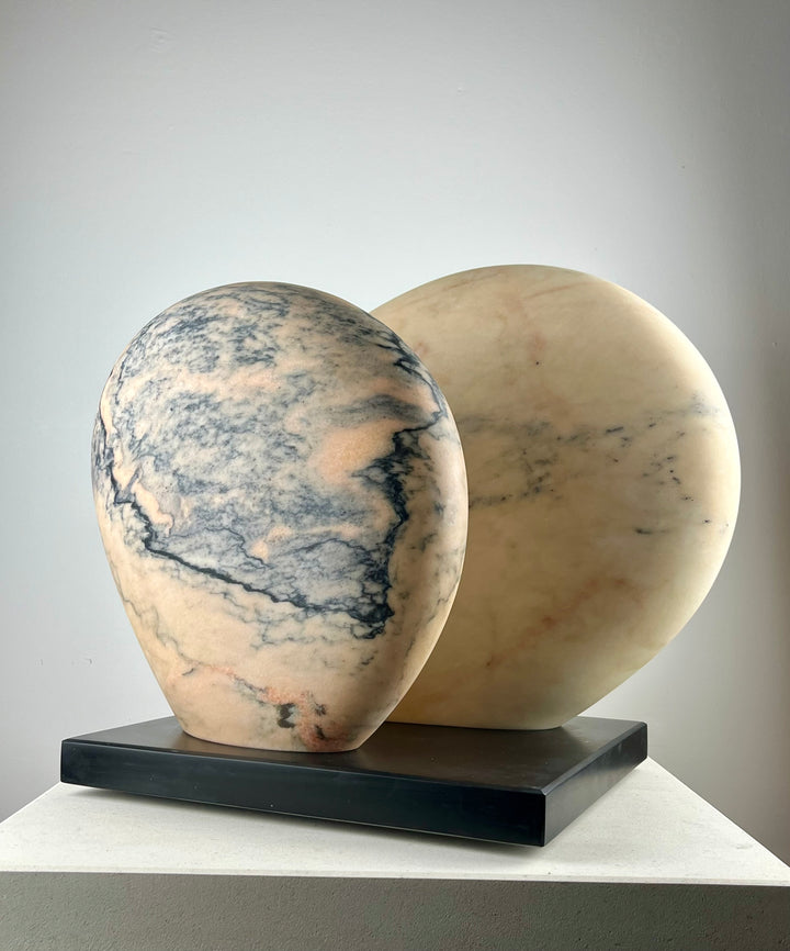 Jason Mulligan sculpture titled APRIL MOON, a one-off hand-carved abstract artwork in Rosa Aurora marble. The piece features two softly rounded, interlocking forms with smooth polished surfaces, revealing delicate tones of pale rose, peach, cream and grey-blue veining. Resting on a dark plinth, the sculpture evokes themes of lunar cycles, balance and renewal, blending timeless marble carving traditions with a contemporary minimalist aesthetic.