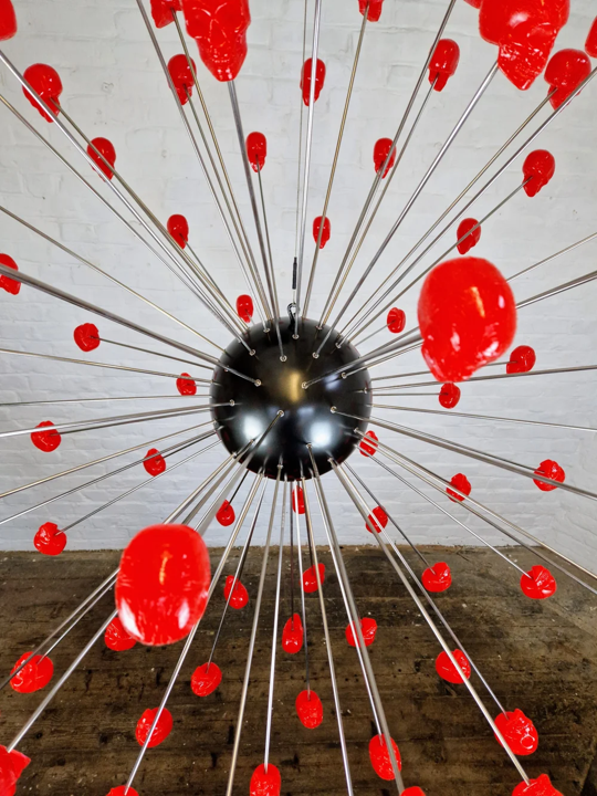 Large spherical hanging sculpture composed of a central black metallic orb with over 70 radiating stainless steel rods, each tipped with a glossy red sculpted skull. The skulls appear to orbit the sphere like a molecular explosion or stylised dandelion, creating a dramatic and surreal visual impact. Materials include stainless steel, polymorph and resin, with vibrant red gloss details and industrial metallic finishes.