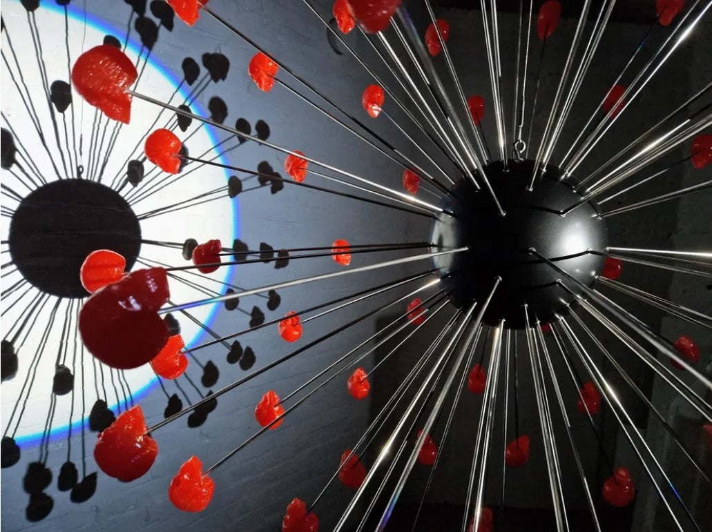 Large spherical hanging sculpture composed of a central black metallic orb with over 70 radiating stainless steel rods, each tipped with a glossy red sculpted skull. The skulls appear to orbit the sphere like a molecular explosion or stylised dandelion, creating a dramatic and surreal visual impact. Materials include stainless steel, polymorph and resin, with vibrant red gloss details and industrial metallic finishes.