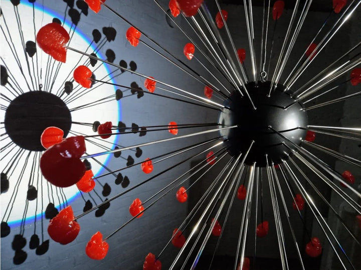 Large spherical hanging sculpture composed of a central black metallic orb with over 70 radiating stainless steel rods, each tipped with a glossy red sculpted skull. The skulls appear to orbit the sphere like a molecular explosion or stylised dandelion, creating a dramatic and surreal visual impact. Materials include stainless steel, polymorph and resin, with vibrant red gloss details and industrial metallic finishes.