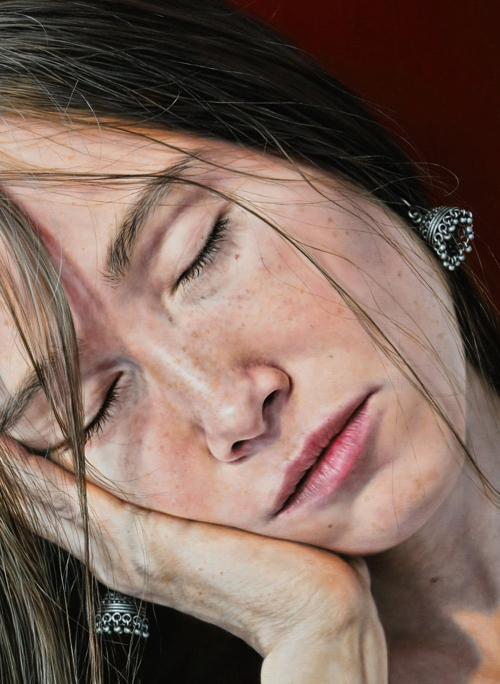 Arina Gordienko hyperrealist oil painting I Had a Dream, portraying a young woman with closed eyes and long brown hair falling across her face, resting her head in her hand, wearing a black top and silver earrings, against a deep red background. The work captures fragility, exhaustion, and dreamlike escape with extraordinary lifelike detail.