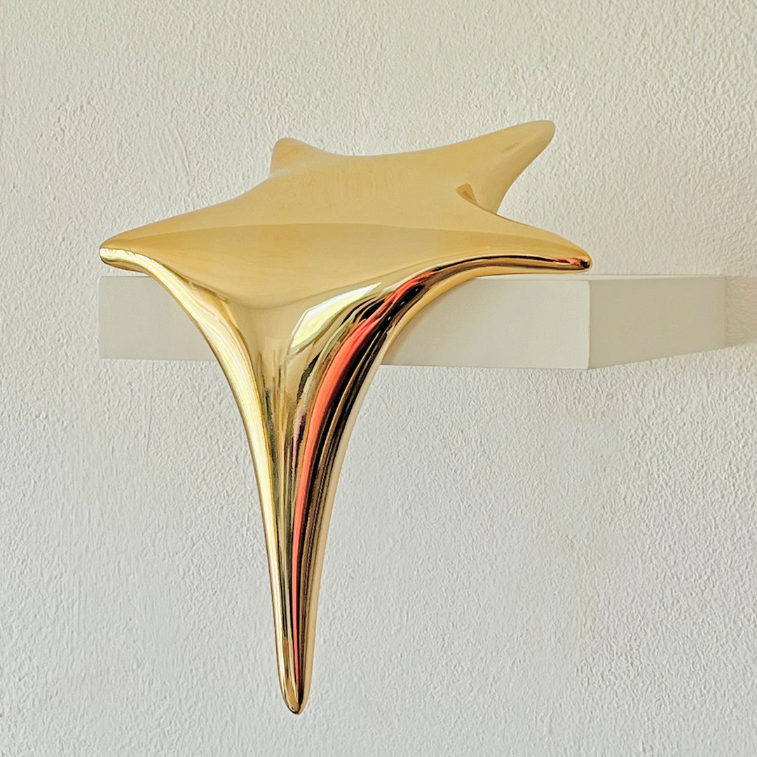 Polished gold-toned stainless steel sculpture in the form of a stylised shooting star, with a fluid, elongated spike melting over the edge of a white plinth or shelf.