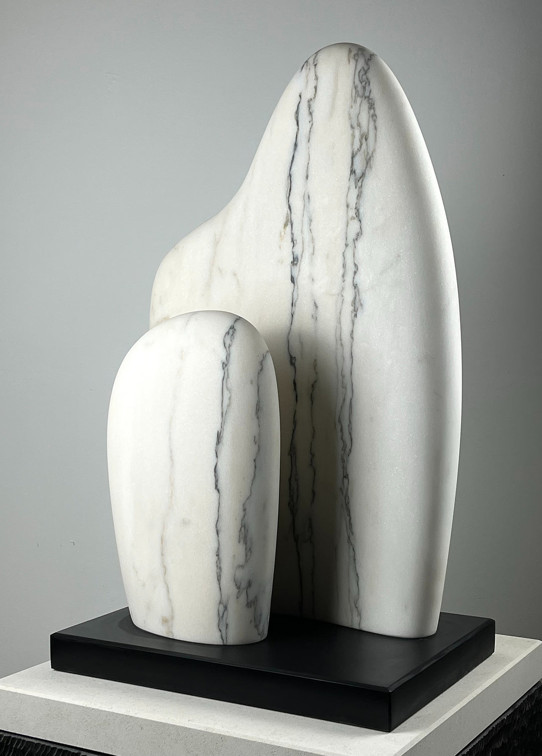 Jason Mulligan sculpture titled LINEAGE, a one-off hand-carved abstract artwork in Portuguese marble. The sculpture features two upright interlocking forms, one taller and one smaller, placed closely together to suggest themes of ancestry, continuity and protection. The smooth surfaces are accented with delicate grey veining running vertically through the creamy white marble, creating a refined balance of strength and tenderness. Resting on a dark plinth, the piece embodies Mulligan’s signature style.