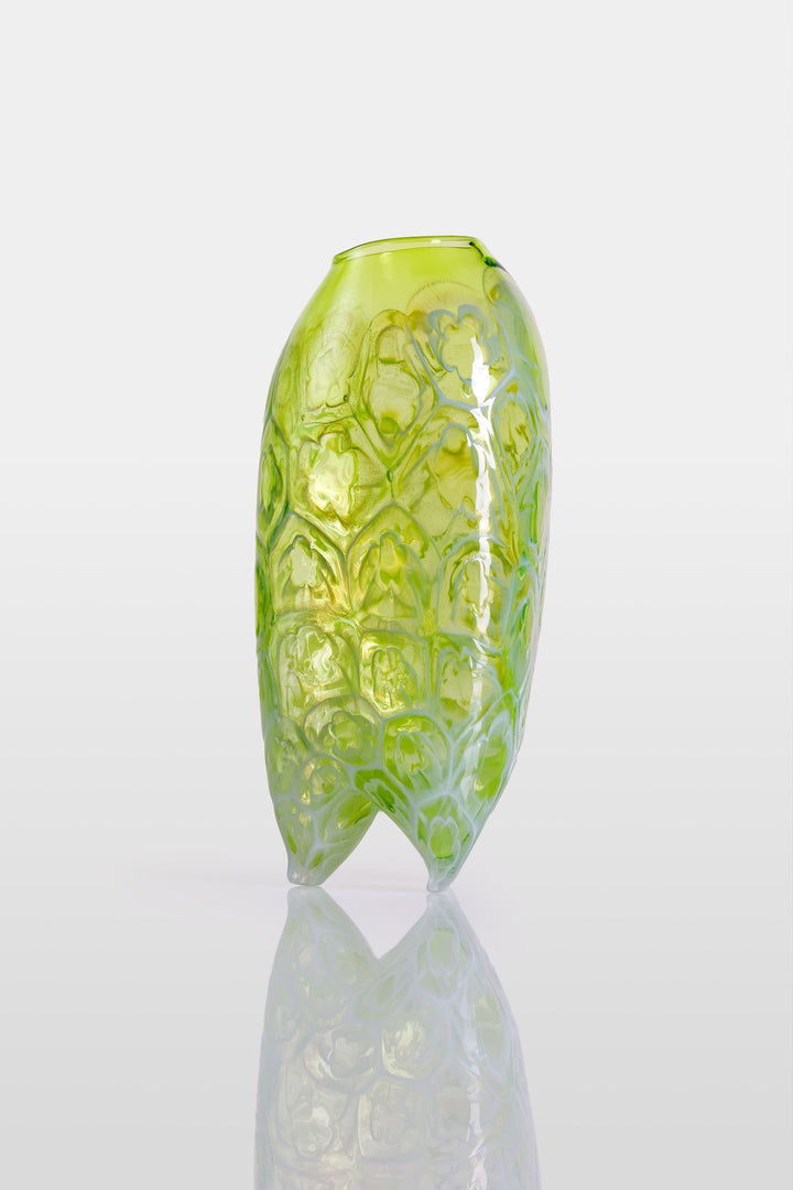 Tall handblown green glass sculpture with embossed floral motifs and a softly asymmetrical, pod-like form, resting on a reflective surface.
