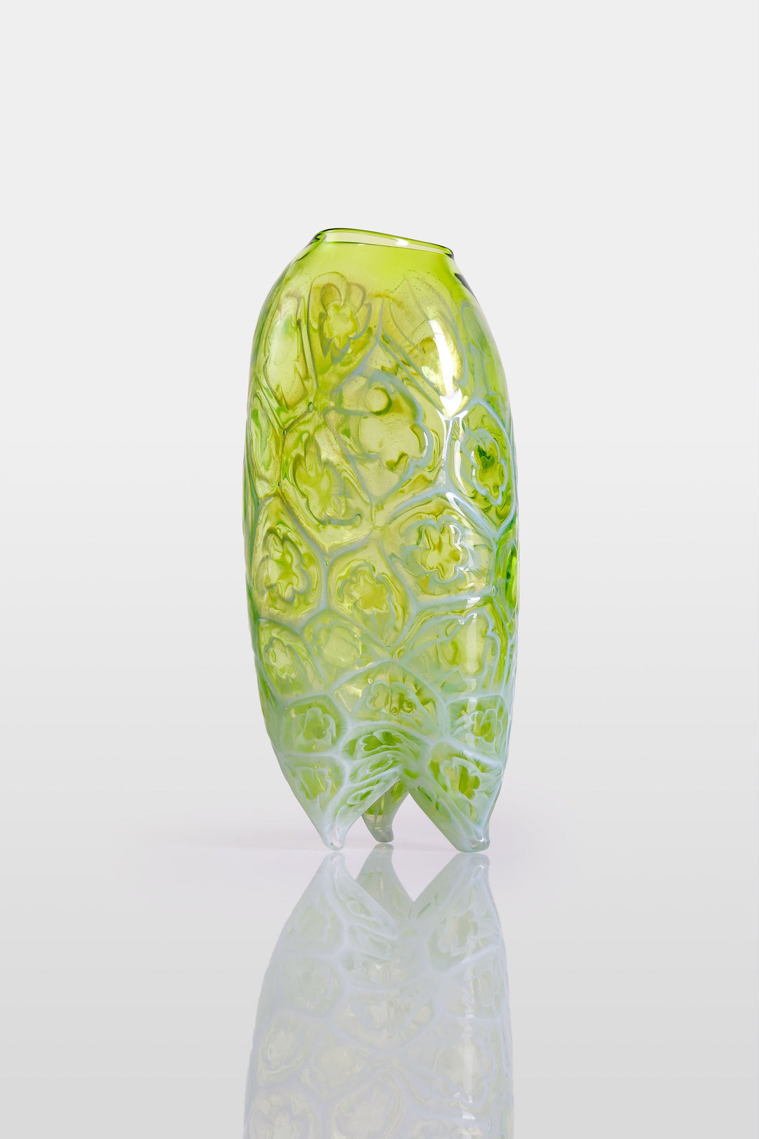 Tall handblown green glass sculpture with embossed floral motifs and a softly asymmetrical, pod-like form, resting on a reflective surface.