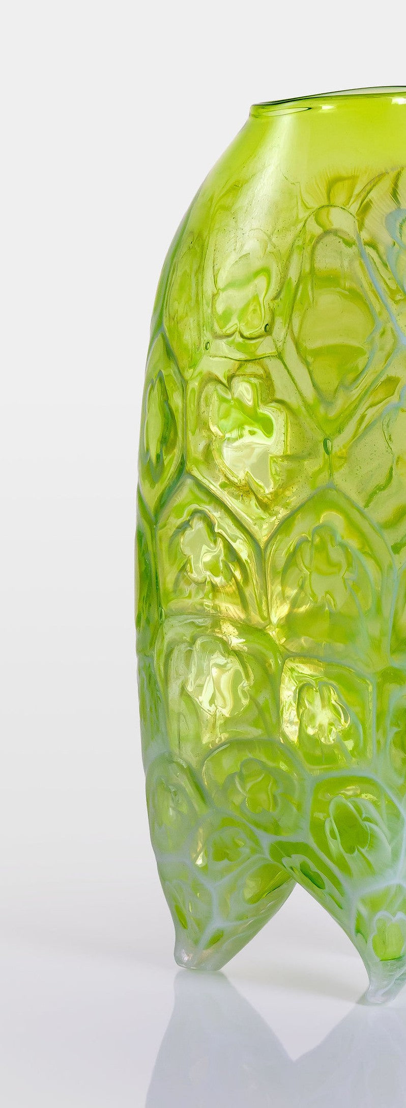 Tall handblown green glass sculpture with embossed floral motifs and a softly asymmetrical, pod-like form, resting on a reflective surface.