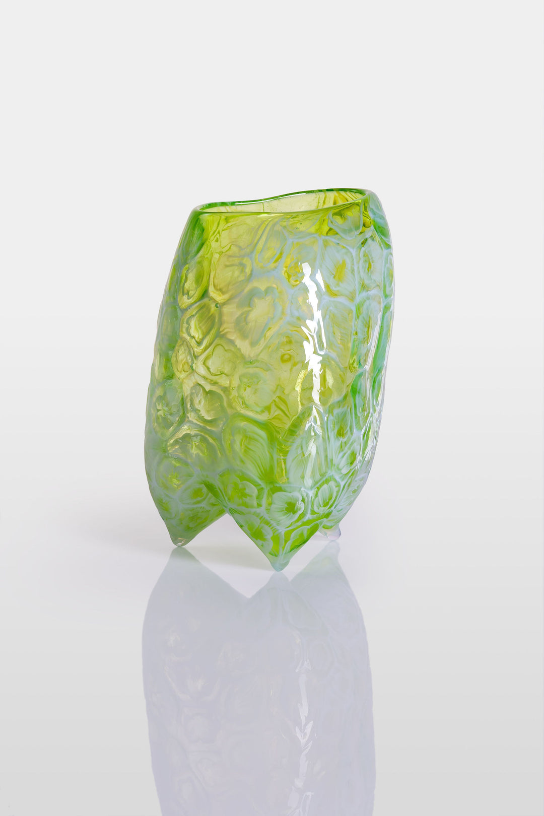 Handcrafted green glass vessel with organic floral embossing and a softly asymmetrical form, set against a white background with reflective base.