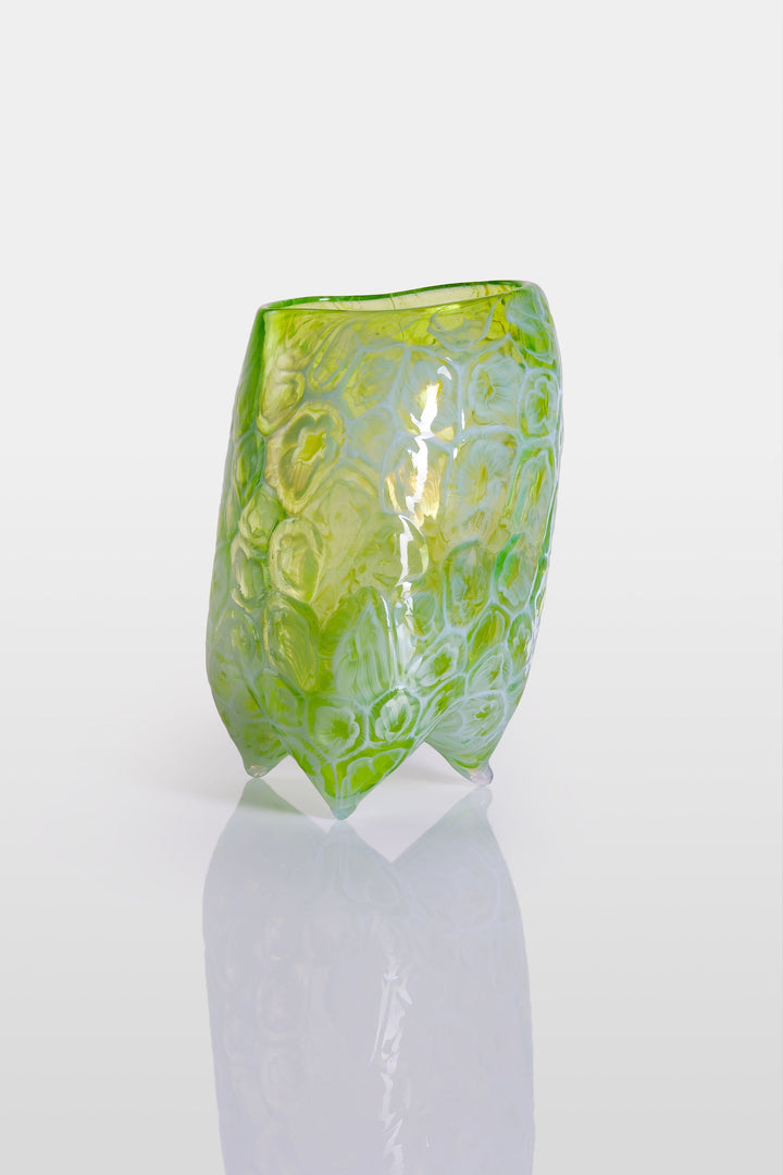 Handcrafted green glass vessel with organic floral embossing and a softly asymmetrical form, set against a white background with reflective base.