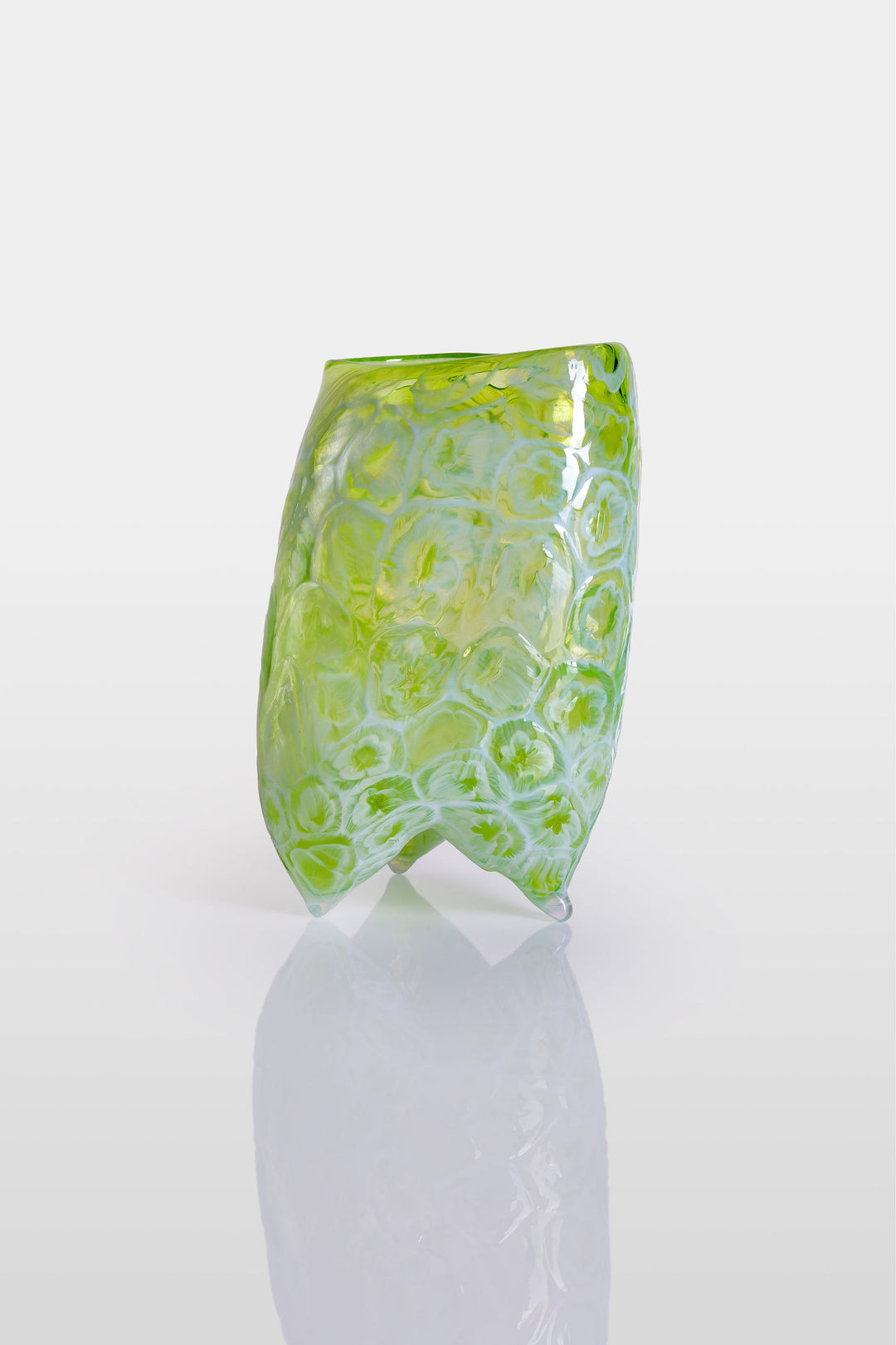 Handcrafted green glass vessel with organic floral embossing and a softly asymmetrical form, set against a white background with reflective base.