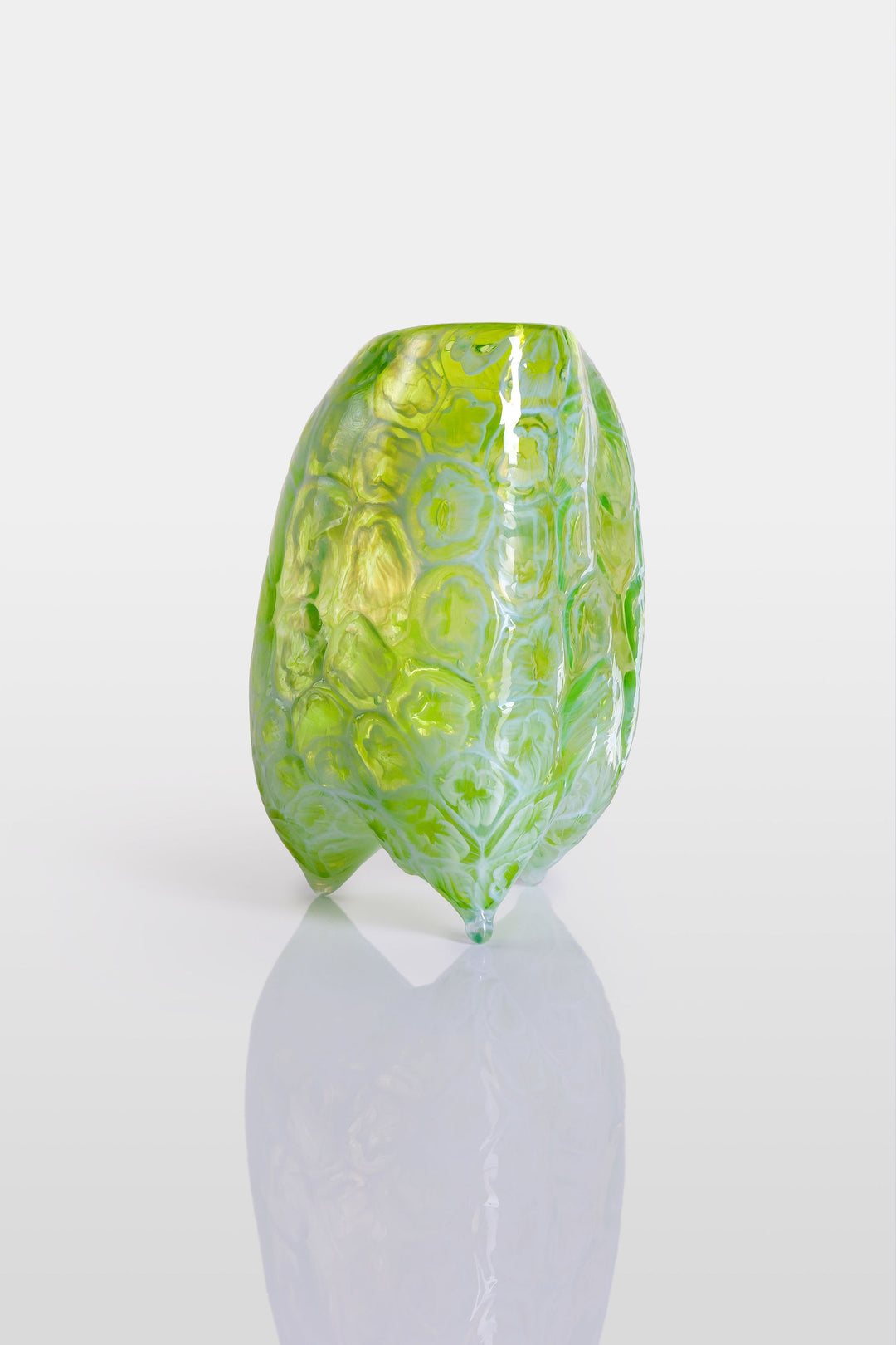 Handcrafted green glass vessel with organic floral embossing and a softly asymmetrical form, set against a white background with reflective base.
