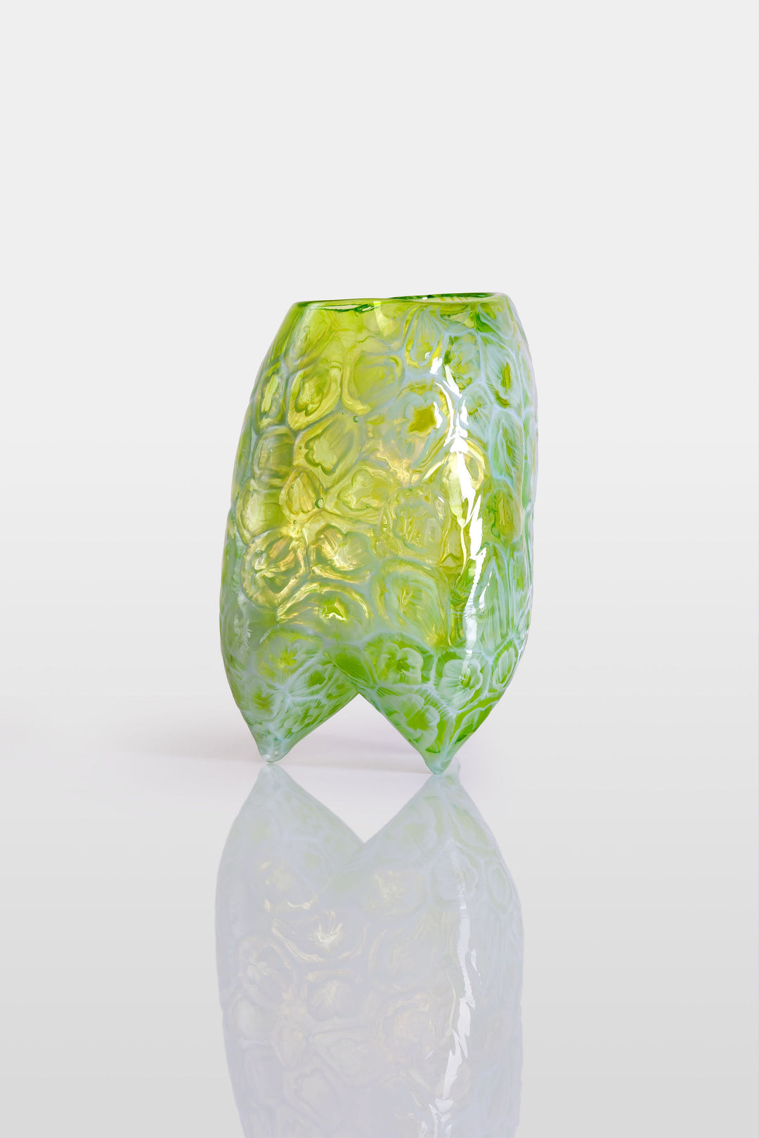 Handcrafted green glass vessel with organic floral embossing and a softly asymmetrical form, set against a white background with reflective base.