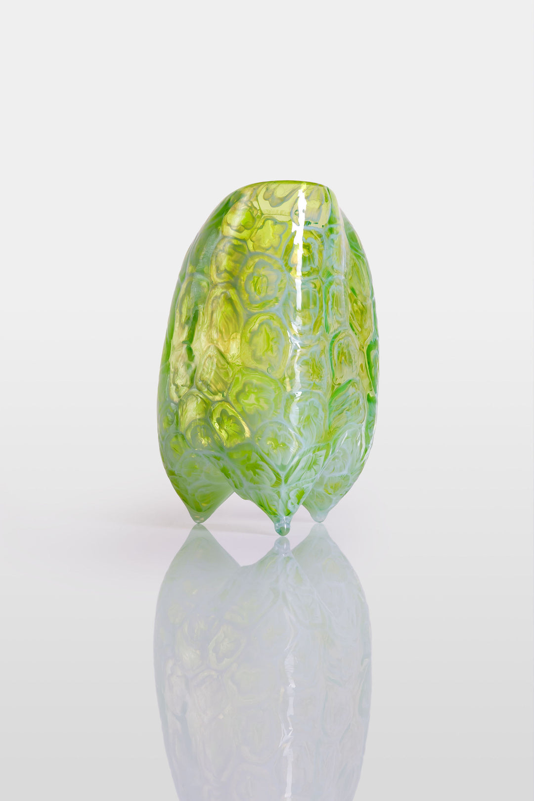 Handcrafted green glass vessel with organic floral embossing and a softly asymmetrical form, set against a white background with reflective base.