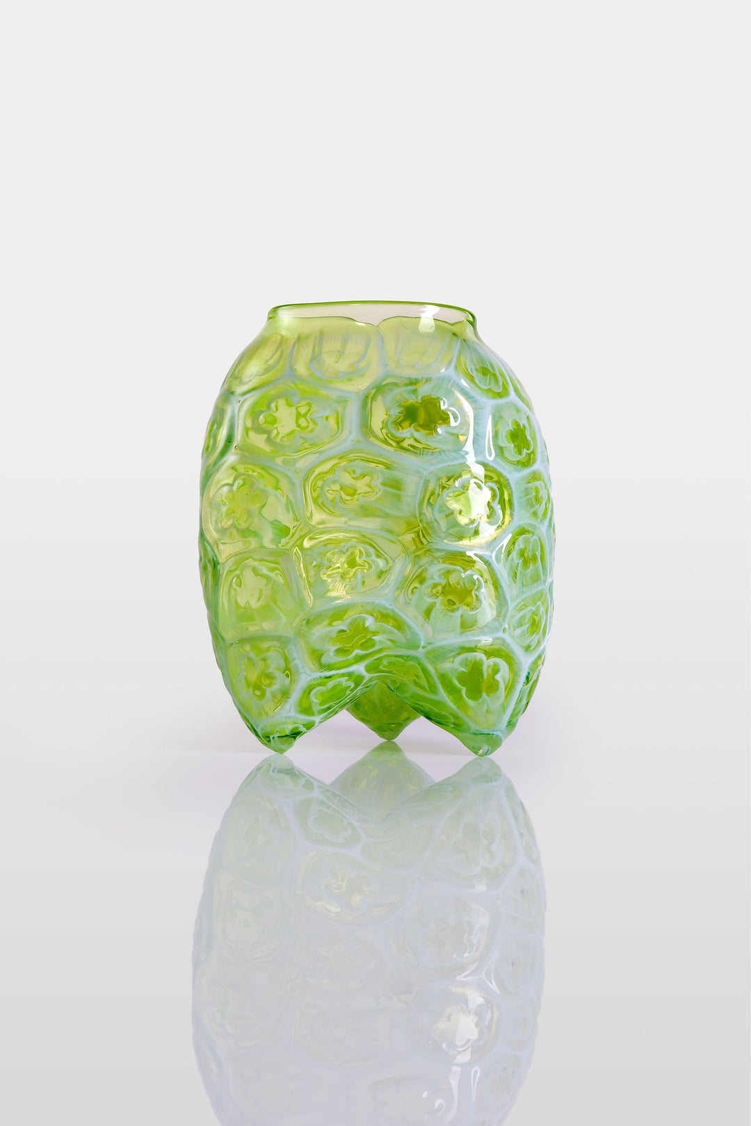 Green hand-blown glass vessel featuring a textured surface with embossed floral motifs and a gently pinched base, shown against a white reflective background.