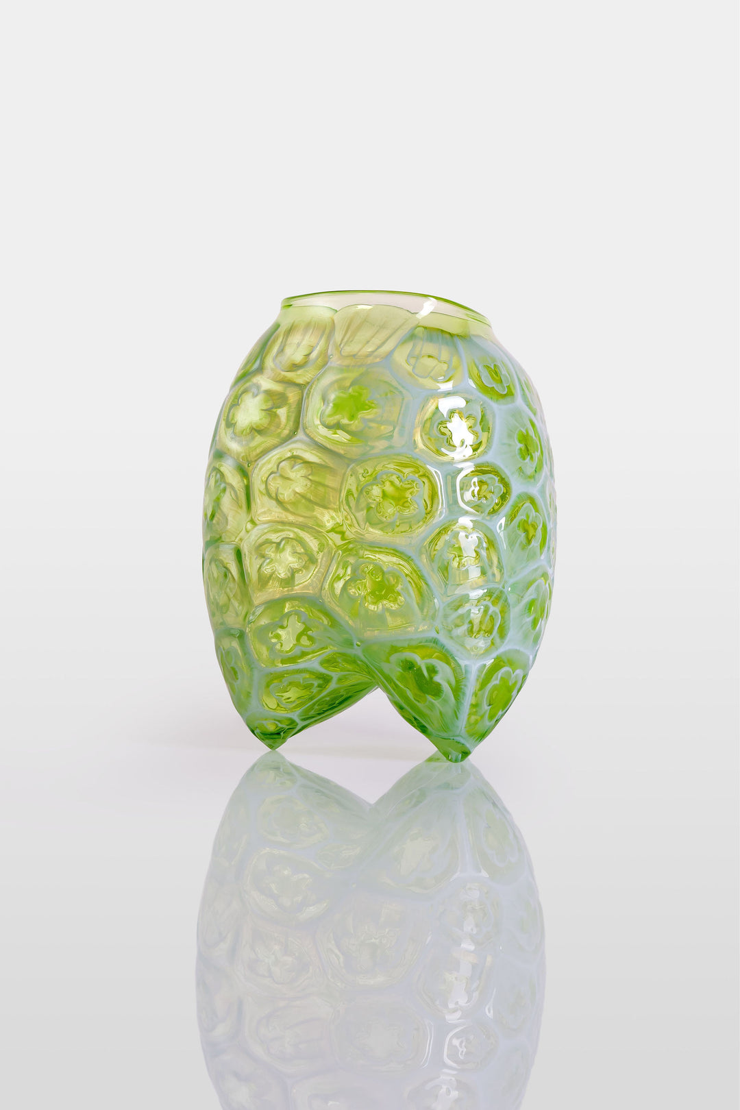 Green hand-blown glass vessel featuring a textured surface with embossed floral motifs and a gently pinched base, shown against a white reflective background.