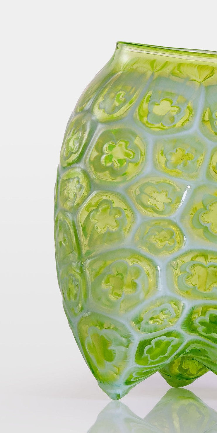 Green hand-blown glass vessel featuring a textured surface with embossed floral motifs and a gently pinched base, shown against a white reflective background.