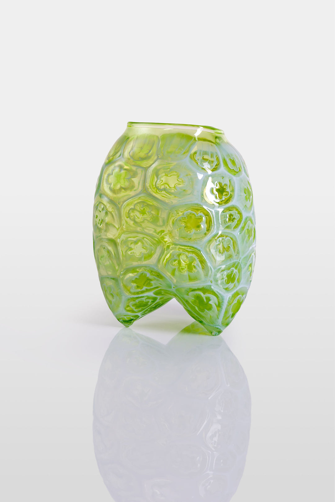 Green hand-blown glass vessel featuring a textured surface with embossed floral motifs and a gently pinched base, shown against a white reflective background.