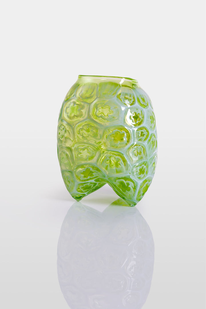 Green hand-blown glass vessel featuring a textured surface with embossed floral motifs and a gently pinched base, shown against a white reflective background.