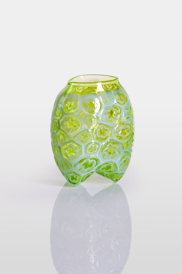 Green hand-blown glass vessel featuring a textured surface with embossed floral motifs and a gently pinched base, shown against a white reflective background.