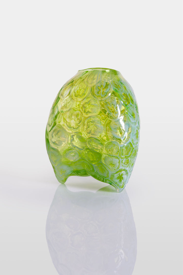 Hand-blown green glass vessel with an organic, asymmetric form and embossed floral patterns, resting on a reflective white surface.