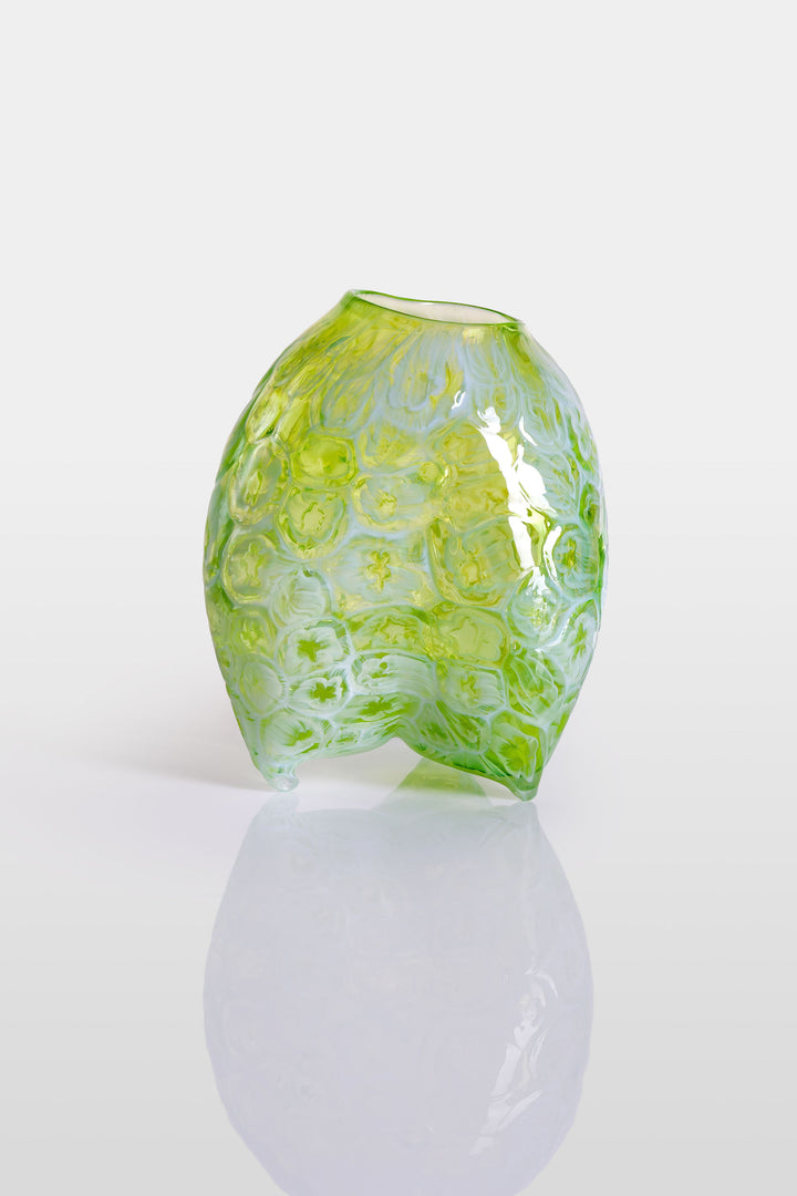 Hand-blown green glass vessel with an organic, asymmetric form and embossed floral patterns, resting on a reflective white surface.