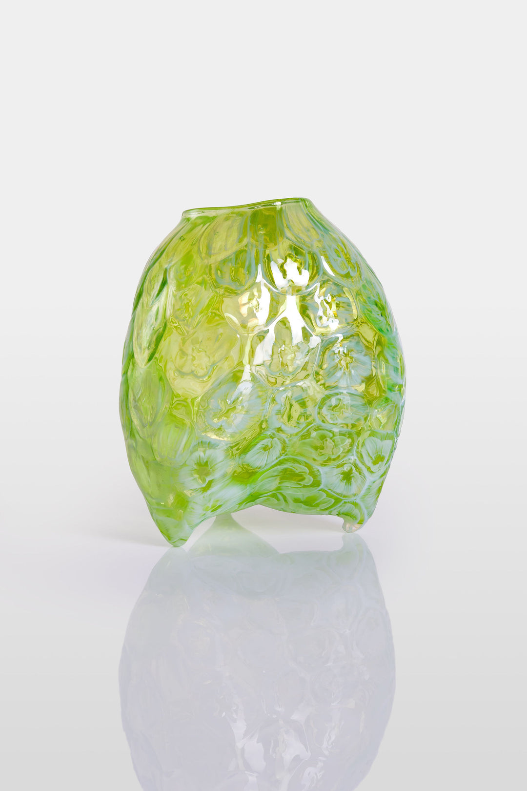 Hand-blown green glass vessel with an organic, asymmetric form and embossed floral patterns, resting on a reflective white surface.