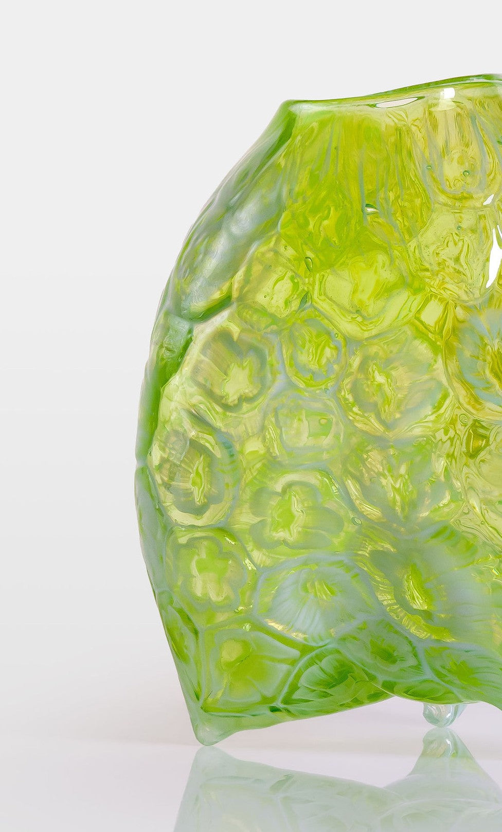 Hand-blown green glass vessel with an organic, asymmetric form and embossed floral patterns, resting on a reflective white surface.