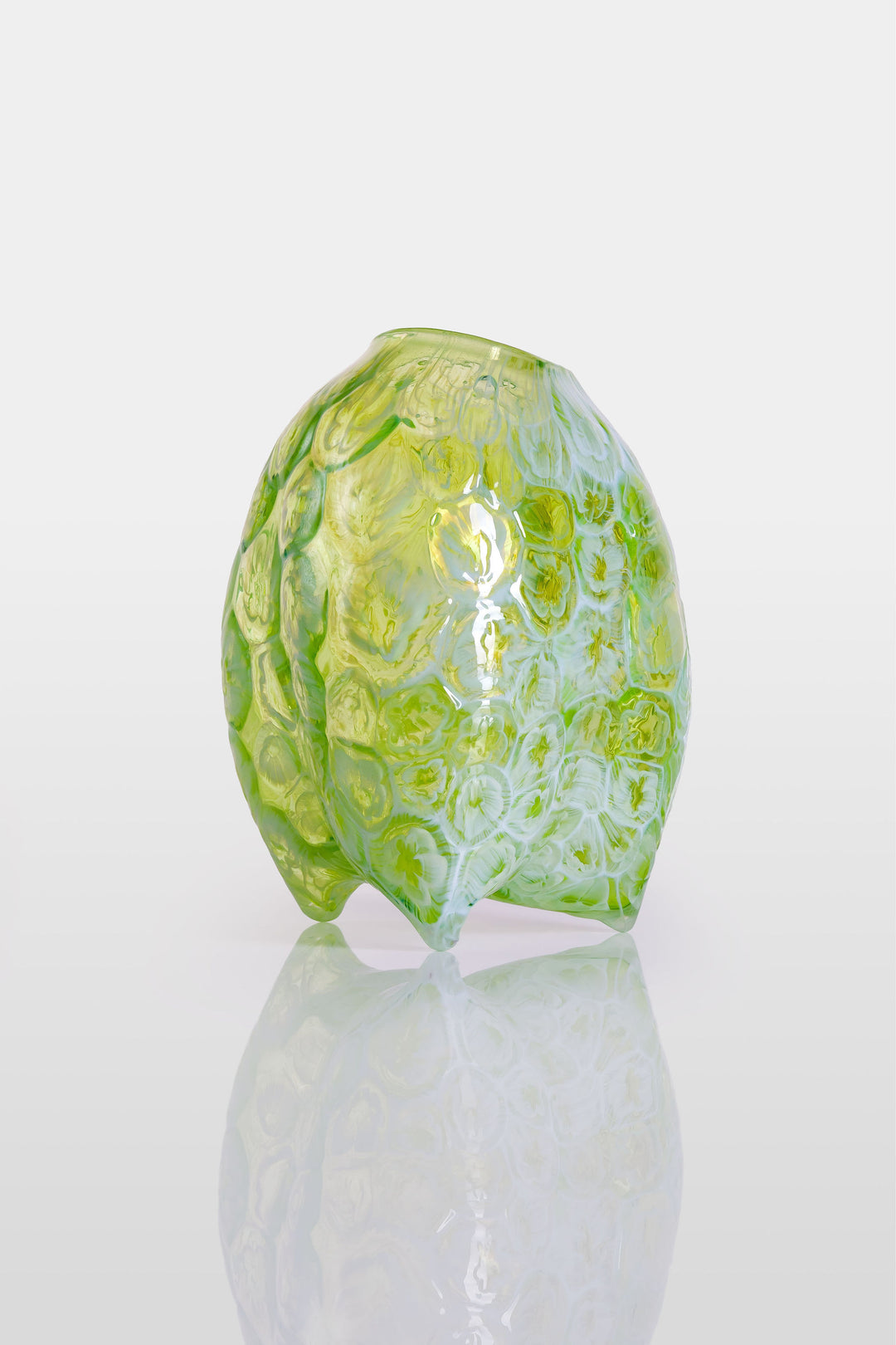 Hand-blown green glass vessel with an organic, asymmetric form and embossed floral patterns, resting on a reflective white surface.
