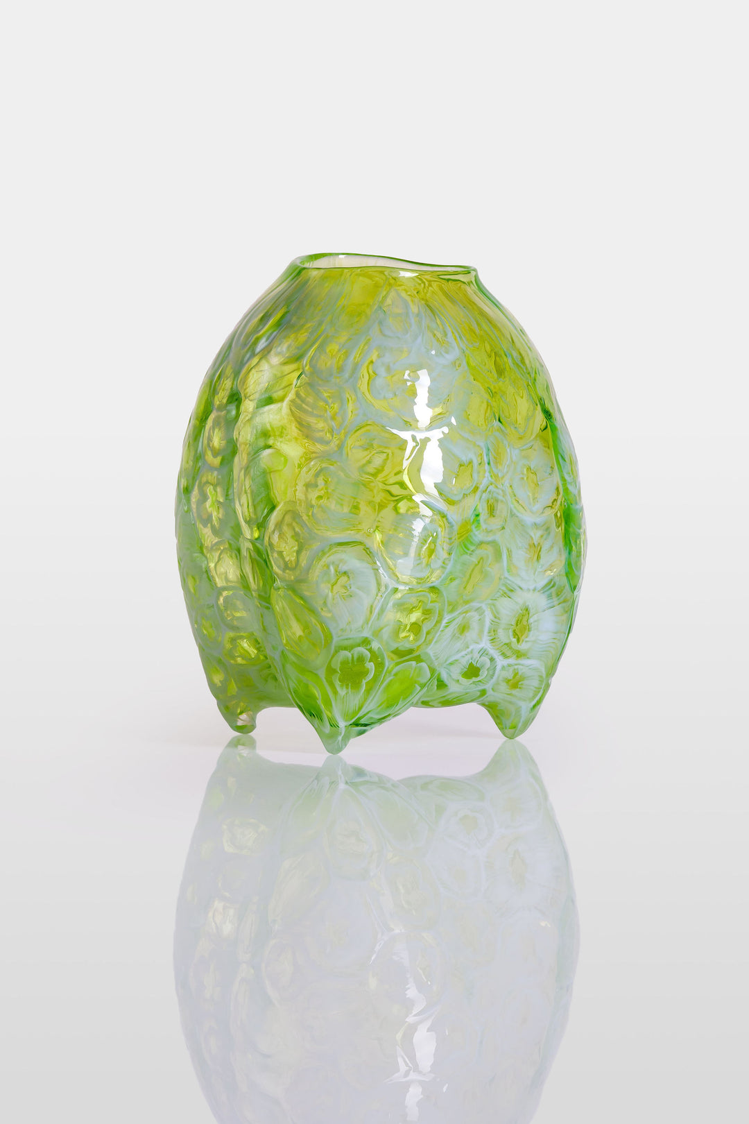 Hand-blown green glass vessel with an organic, asymmetric form and embossed floral patterns, resting on a reflective white surface.