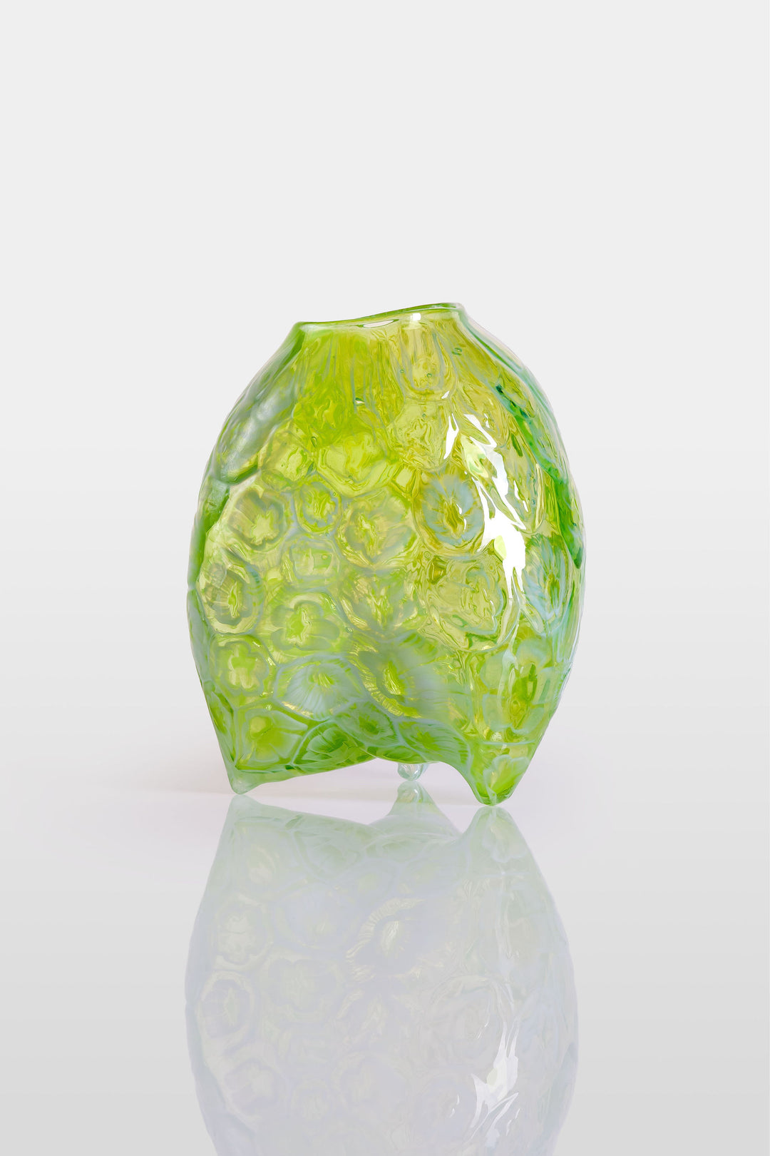 Hand-blown green glass vessel with an organic, asymmetric form and embossed floral patterns, resting on a reflective white surface.