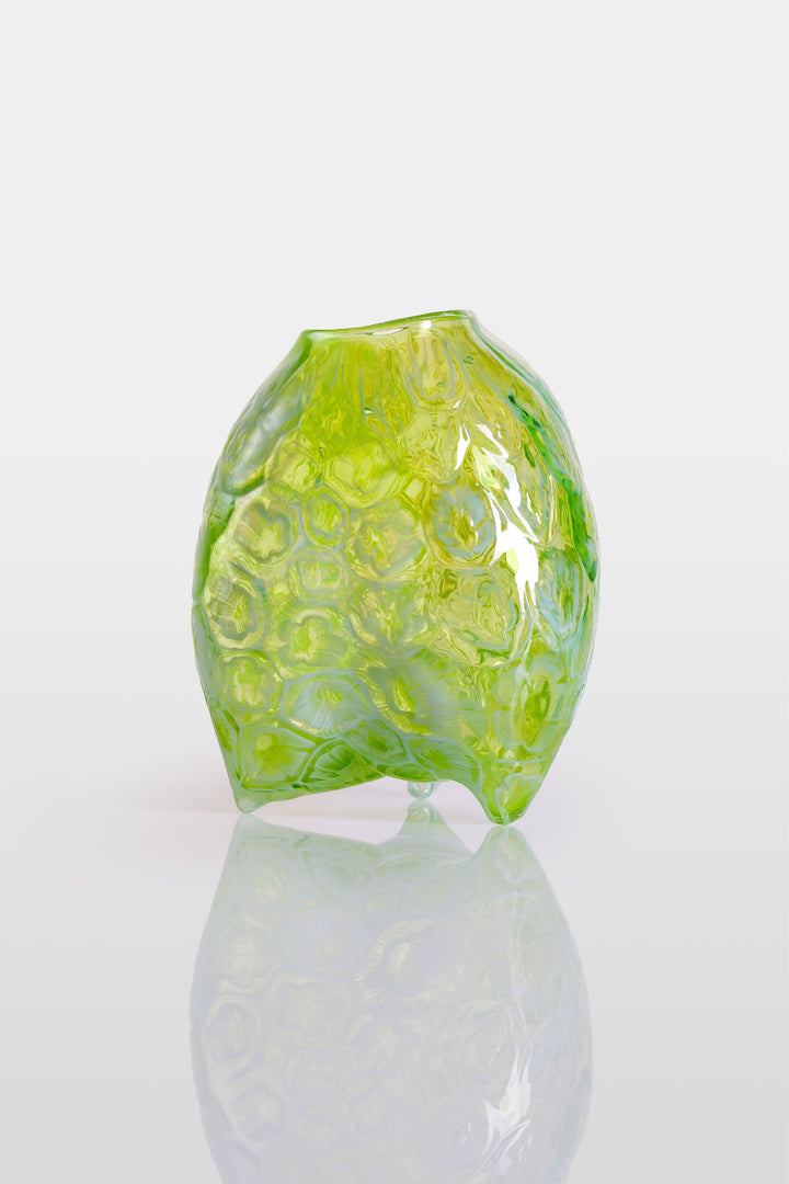 Hand-blown green glass vessel with an organic, asymmetric form and embossed floral patterns, resting on a reflective white surface.