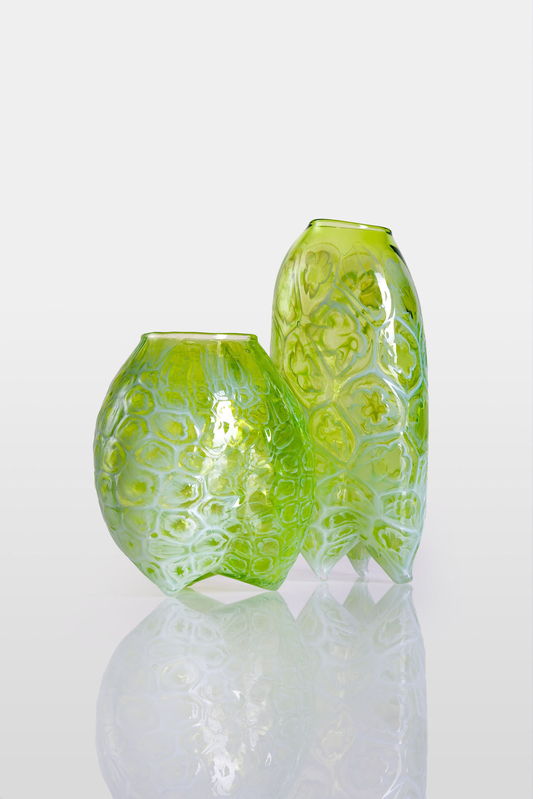 Tall handblown green glass sculpture with embossed floral motifs and a softly asymmetrical, pod-like form, resting on a reflective surface.