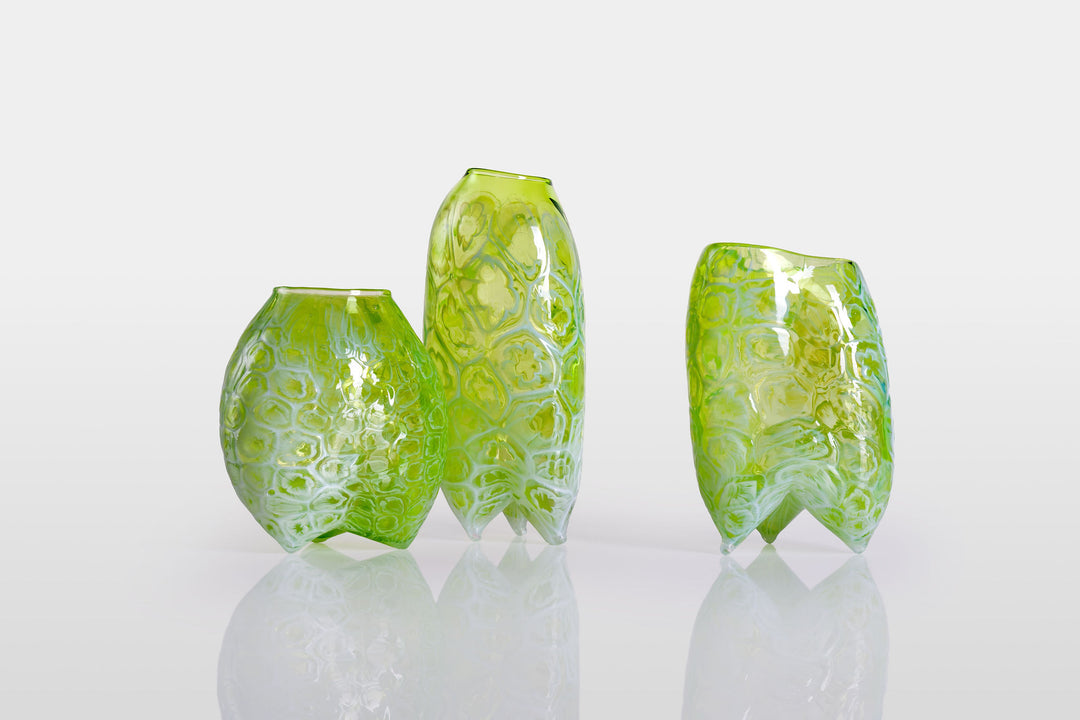 Tall handblown green glass sculpture with embossed floral motifs and a softly asymmetrical, pod-like form, resting on a reflective surface.
