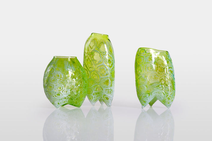 Tall handblown green glass sculpture with embossed floral motifs and a softly asymmetrical, pod-like form, resting on a reflective surface.