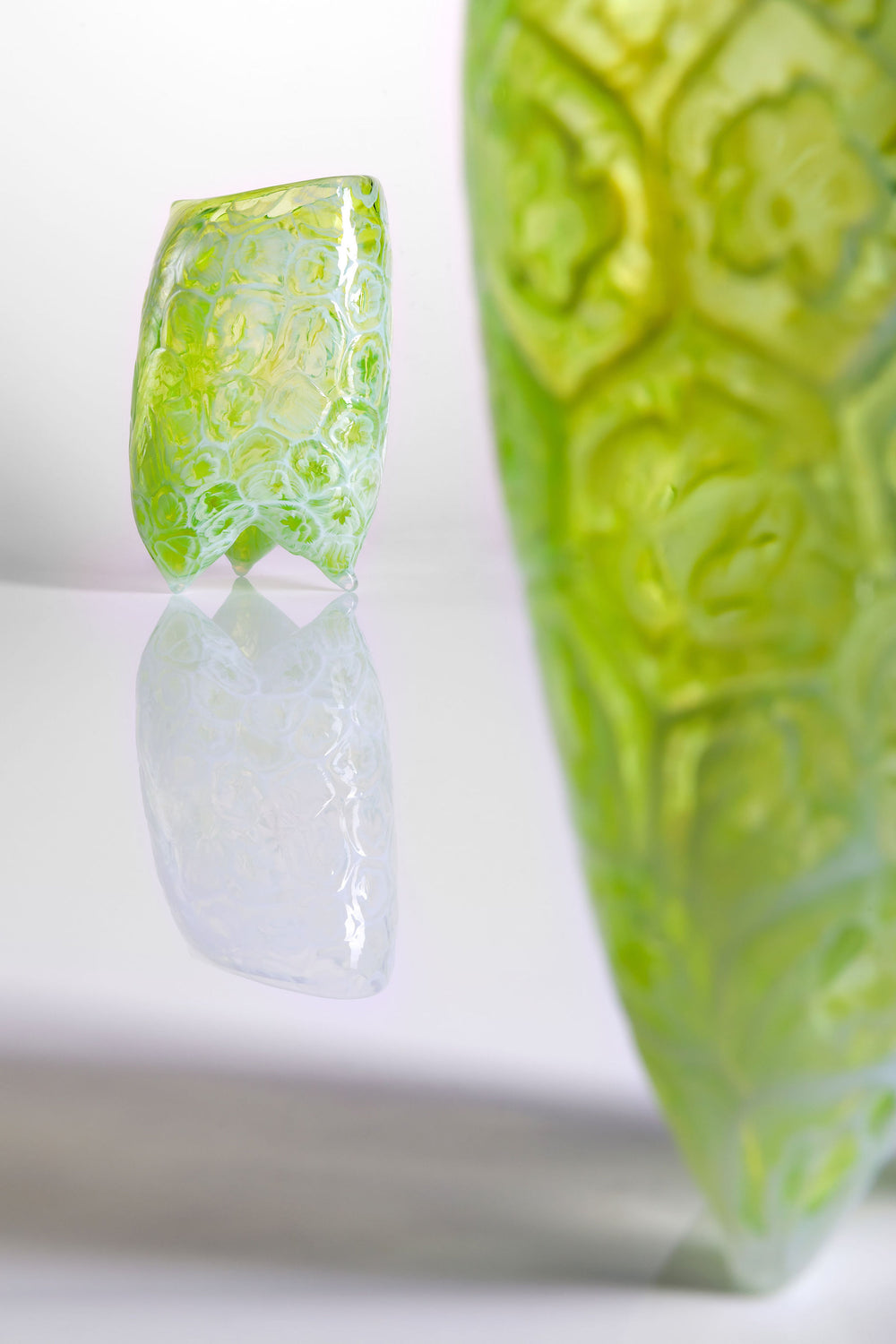 Handcrafted green glass vessel with organic floral embossing and a softly asymmetrical form, set against a white background with reflective base.