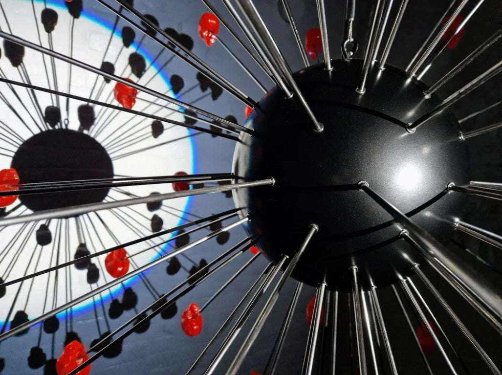 Large spherical hanging sculpture composed of a central black metallic orb with over 70 radiating stainless steel rods, each tipped with a glossy red sculpted skull. The skulls appear to orbit the sphere like a molecular explosion or stylised dandelion, creating a dramatic and surreal visual impact. Materials include stainless steel, polymorph and resin, with vibrant red gloss details and industrial metallic finishes.