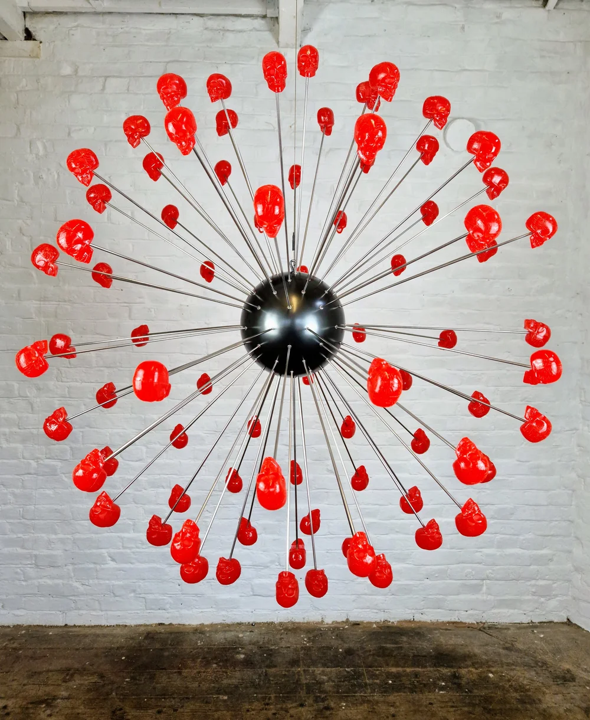Large spherical hanging sculpture composed of a central black metallic orb with over 70 radiating stainless steel rods, each tipped with a glossy red sculpted skull. The skulls appear to orbit the sphere like a molecular explosion or stylised dandelion, creating a dramatic and surreal visual impact. Materials include stainless steel, polymorph and resin, with vibrant red gloss details and industrial metallic finishes.