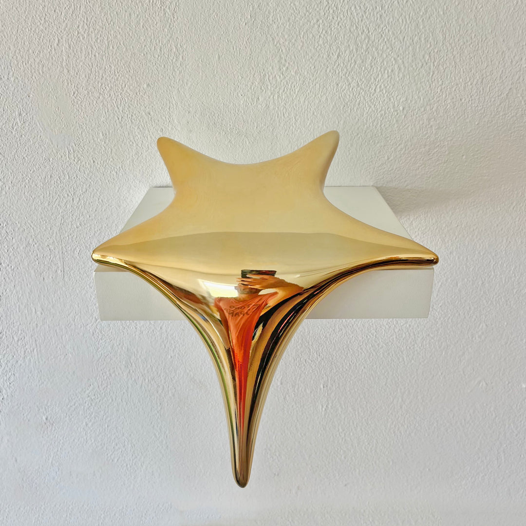 Polished gold-toned stainless steel sculpture in the form of a stylised shooting star, with a fluid, elongated spike melting over the edge of a white plinth or shelf.