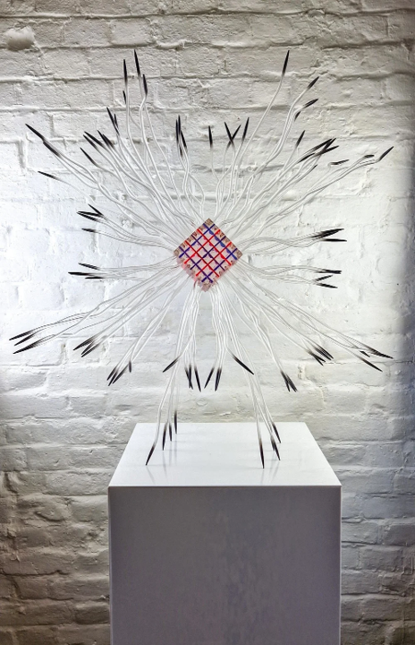 Large-scale sculptural artwork featuring a central geometric cube formed from layered resin in transparent red, blue, and clear tones. Radiating outward are dozens of transparent, sinuous tentacle-like extensions made of acrylic or resin tubing, tipped in black. The structure resembles an imagined extraterrestrial lifeform, with a visually striking symmetry and futuristic energy. Set against a white brick wall, the piece evokes themes of outer space, abstract biology, and the unknown.