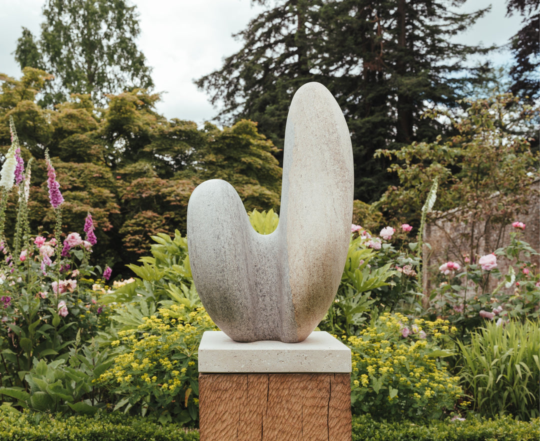 Jason Mulligan sculpture titled LITH, a one-off hand-carved abstract artwork in Ancaster Weatherbed limestone, mounted on an oak base. The organic twin-form rises with a smooth, curving geometry that opens upward, suggesting balance, renewal, and growth. The stone surface has natural earthy tones of soft beige, grey, and warm brown, subtly weathered to highlight the tactile grain of the limestone. Displayed outdoors, the sculpture integrates seamlessly with its natural garden setting.
