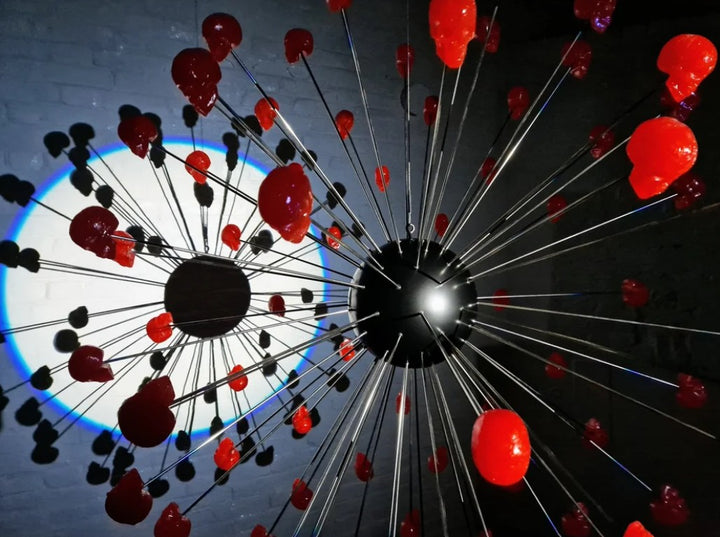 Large spherical hanging sculpture composed of a central black metallic orb with over 70 radiating stainless steel rods, each tipped with a glossy red sculpted skull. The skulls appear to orbit the sphere like a molecular explosion or stylised dandelion, creating a dramatic and surreal visual impact. Materials include stainless steel, polymorph and resin, with vibrant red gloss details and industrial metallic finishes.