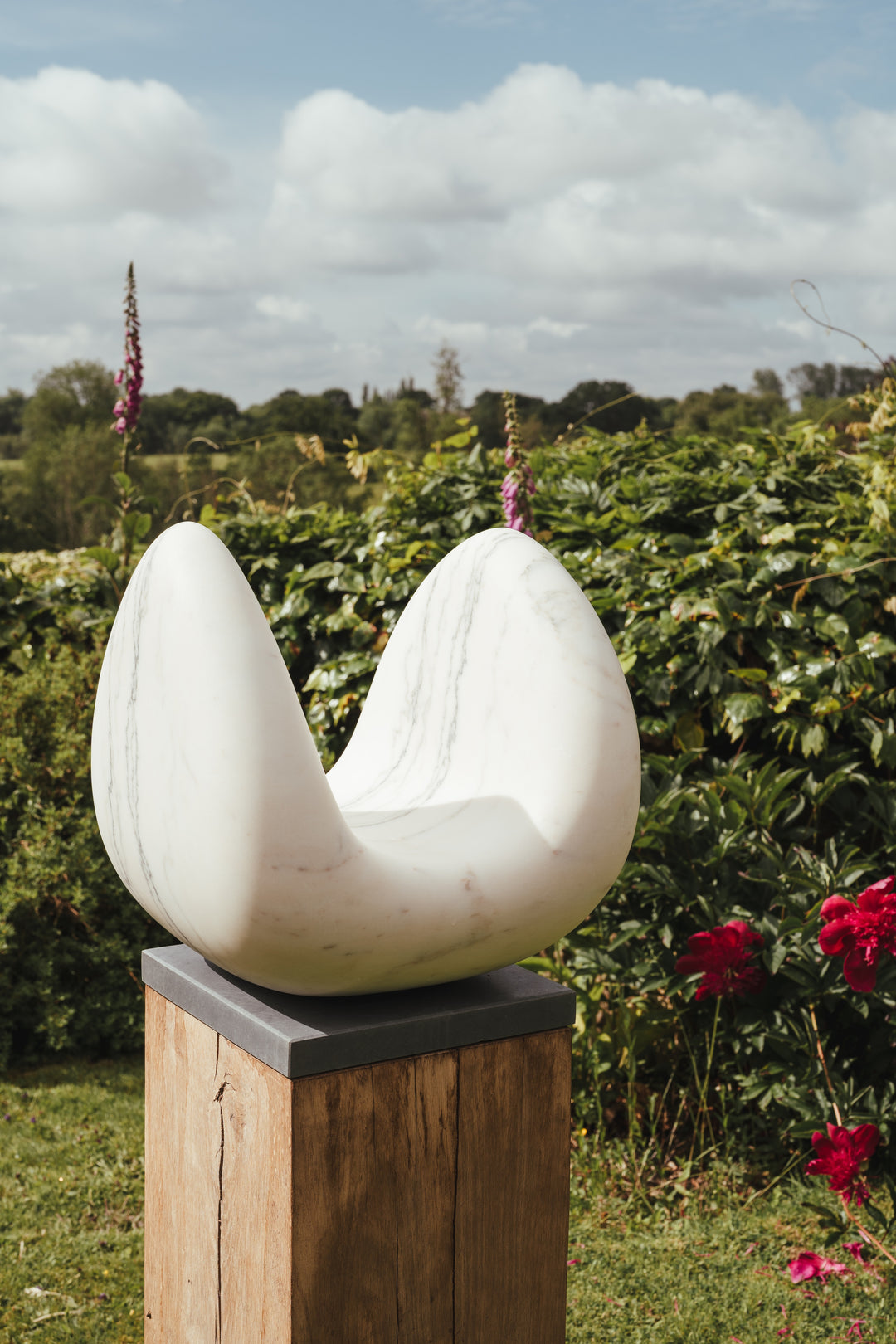 Jason Mulligan sculpture titled PASSAGE, a one-off contemporary abstract work hand-carved from Portuguese marble. The sculpture features an open, twin-peaked form with smooth, curving contours rising from a hollowed centre, suggesting ideas of transition, passage, and flow. The white marble is veined with subtle grey and soft pinkish tones, adding depth and natural variation to the polished surface. Mounted on a tall oak base with a dark stone cap, the piece integrates elegance with strength.