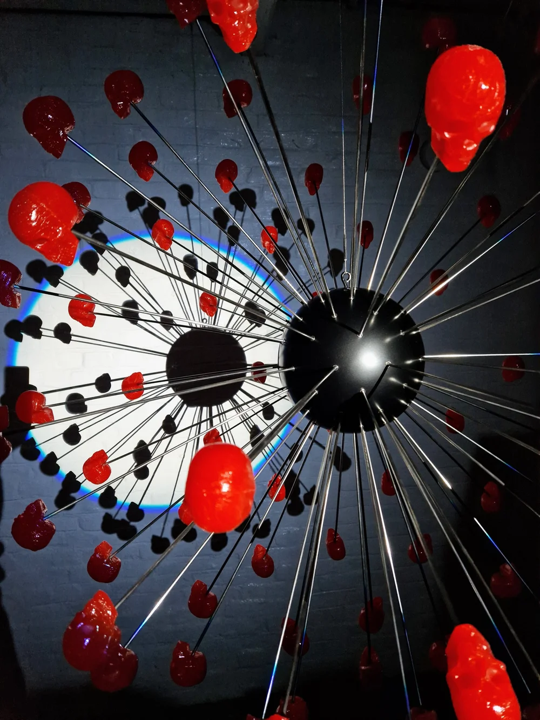 Large spherical hanging sculpture composed of a central black metallic orb with over 70 radiating stainless steel rods, each tipped with a glossy red sculpted skull. The skulls appear to orbit the sphere like a molecular explosion or stylised dandelion, creating a dramatic and surreal visual impact. Materials include stainless steel, polymorph and resin, with vibrant red gloss details and industrial metallic finishes.