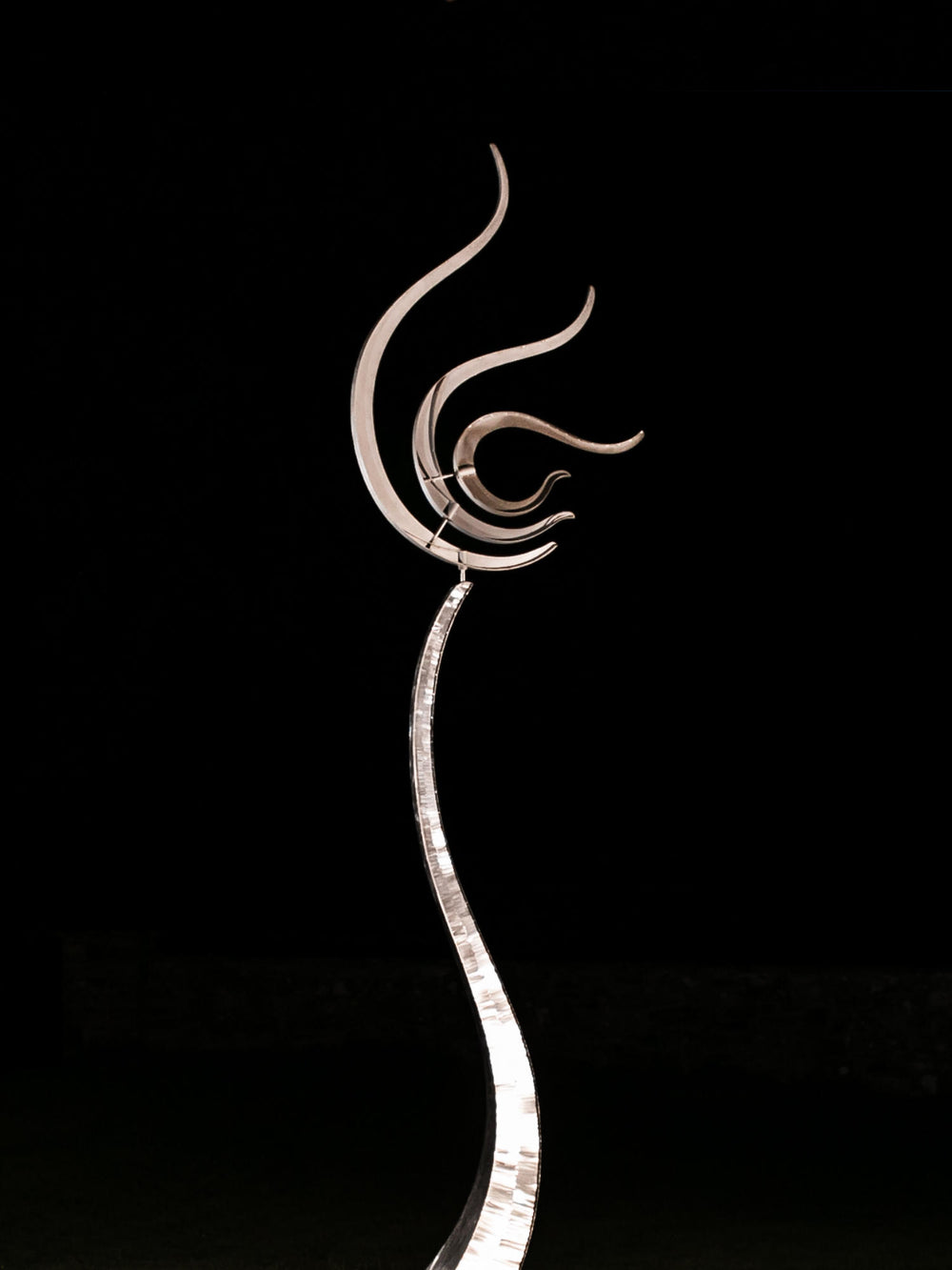Tall stainless steel kinetic sculpture with fluid, flame-like curves, standing against a blue sky in a landscaped garden
