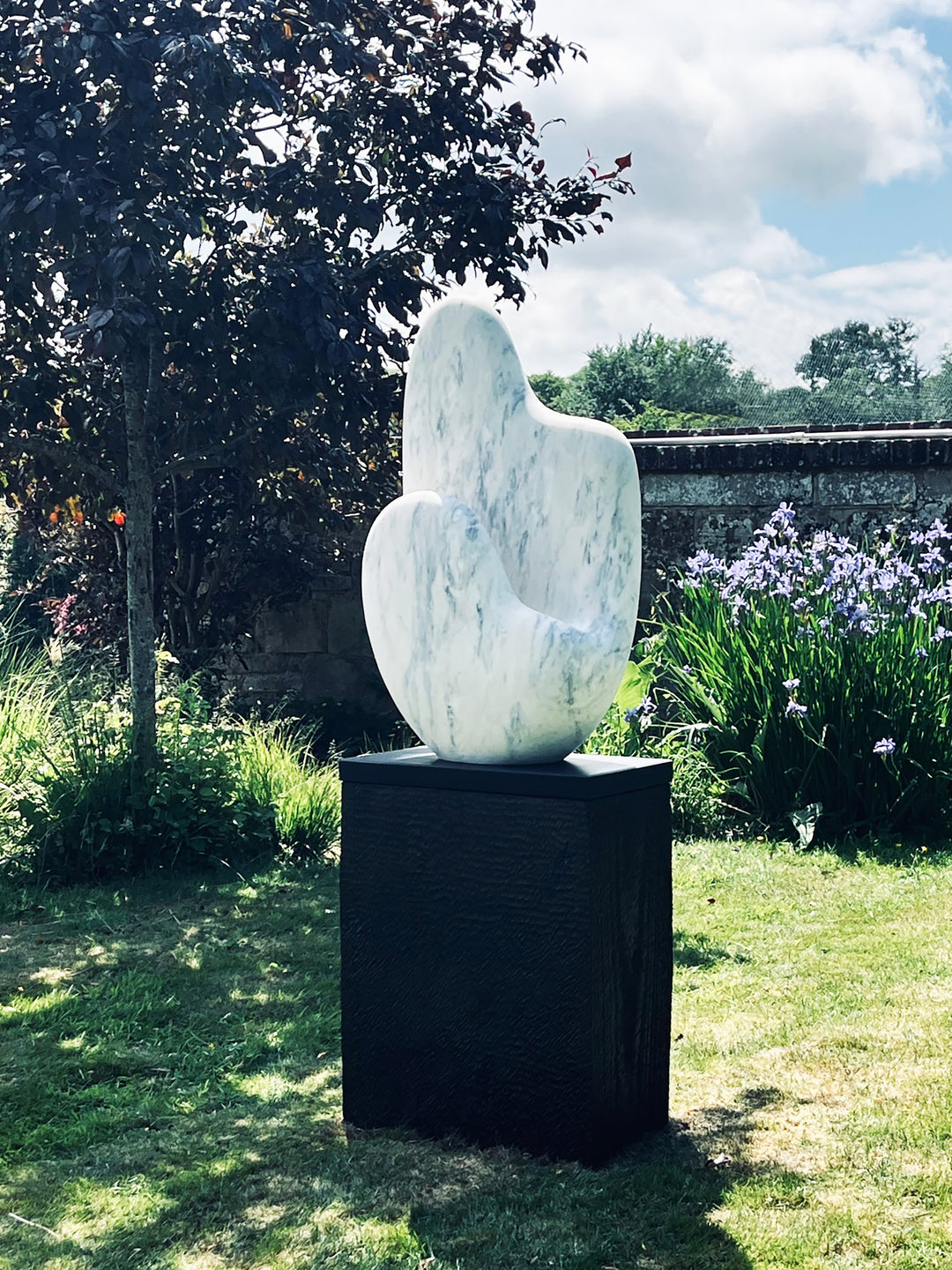 Jason Mulligan sculpture titled KINDRED, a one-off contemporary abstract form carved from Crema Aurora marble. The sculpture features a tall, dual-peaked organic shape with a softly curving hollow, evoking themes of kinship, connection, and balance. The marble surface is smooth and polished, with pale cream tones subtly veined with soft grey and warm blush accents, enhancing its natural elegance. Mounted on a tall dark plinth, the work is designed for both outdoor garden placement and indoor display.